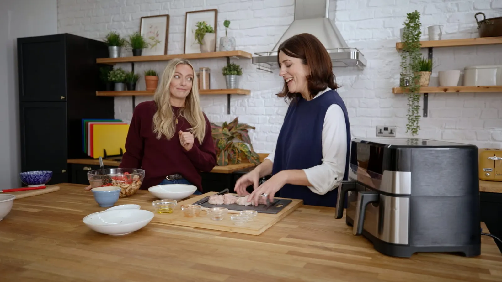 A blonde woman in a maroon jumper watches as a brown-haired woman in a navy top slices meat on a chopping board beside an air fryer.