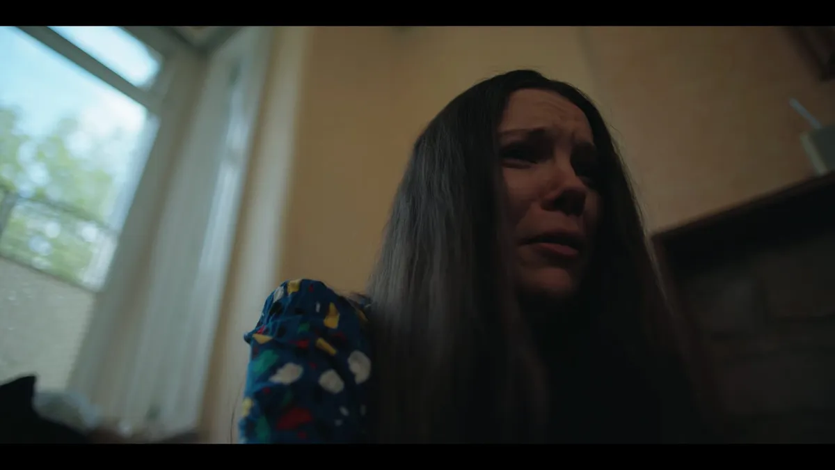 A woman with long dark hair and a patterned top sits by a window, low angle, tearful expression, looking off-camera.