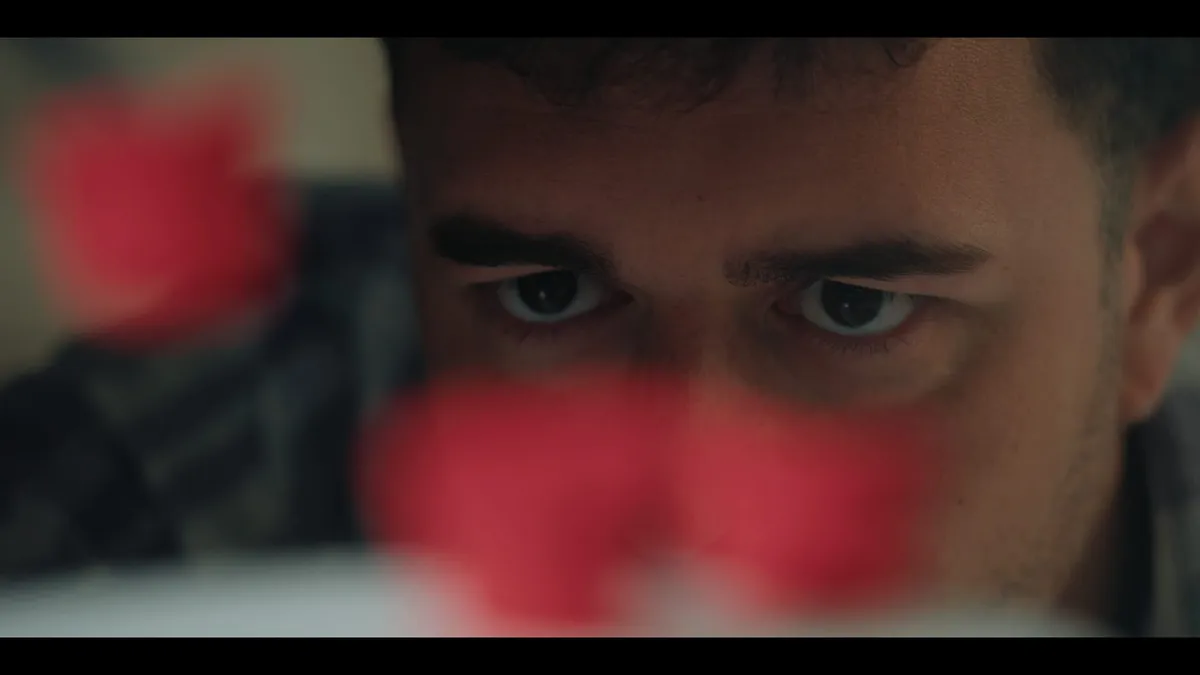 Extreme close-up of a man's eyes and forehead, looking downward, with an out-of-focus red object partially obscuring the lower frame.