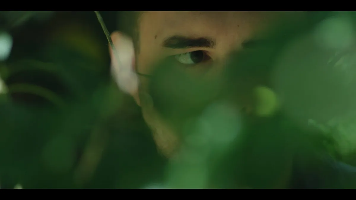 Close-up of a man's eye and brow through foliage, his face partly obscured by blurred green leaves, looking off-camera.