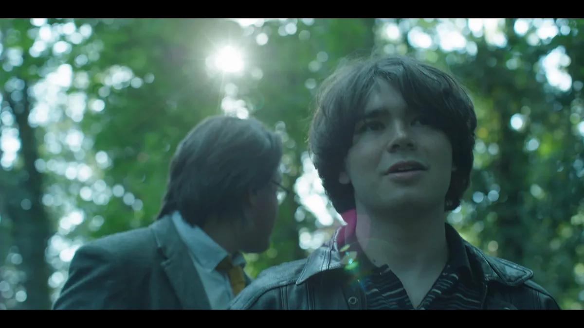 Close-up of a young man with dark hair looking off-camera, an older man in a grey suit beside him, sunlight filtering through trees behind.