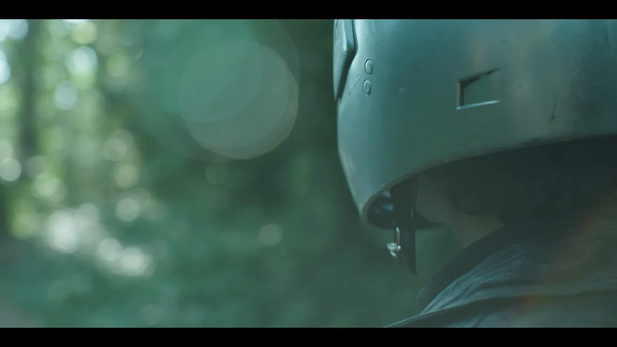 Close-up profile of a person in a military-style helmet with chinstrap, seen from behind against a blurred green woodland background.