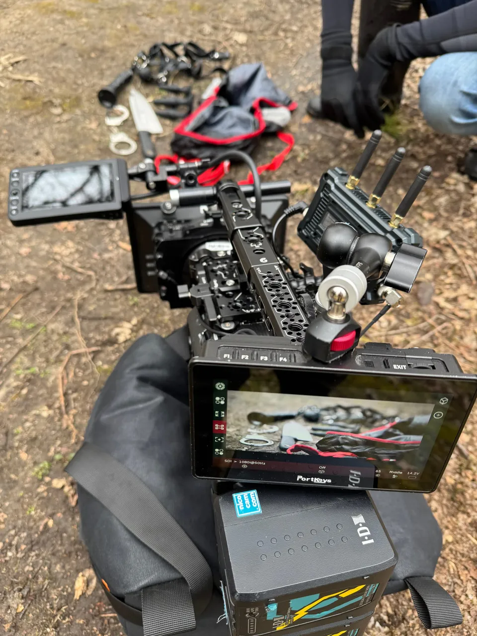 Camera rig with monitor and antennas resting on a bag, set on woodland ground, with a gloved hand and kneeling leg visible behind it.