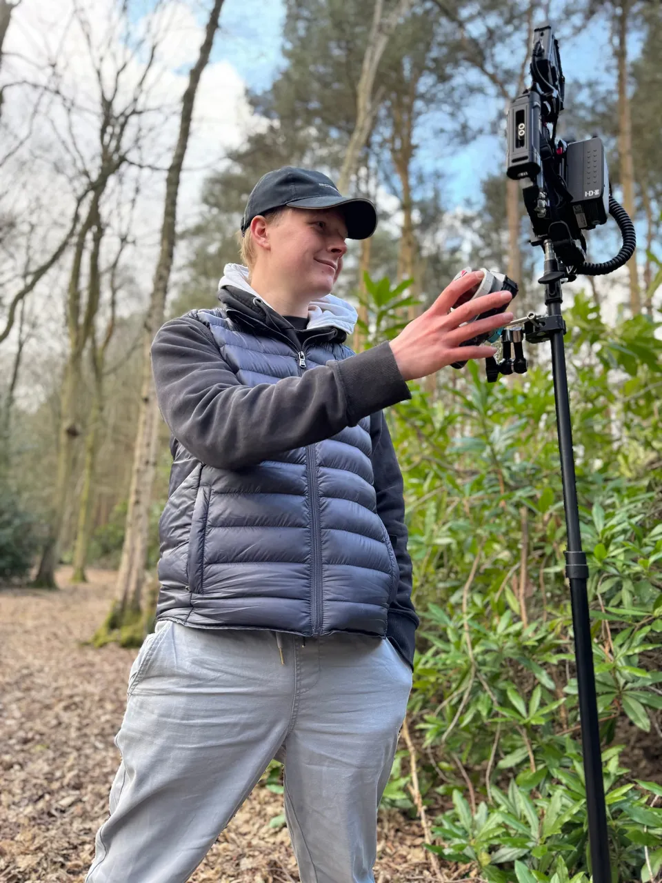 A man in a cap and gilet adjusts equipment mounted on a stand in woodland, surrounded by trees and shrubs.