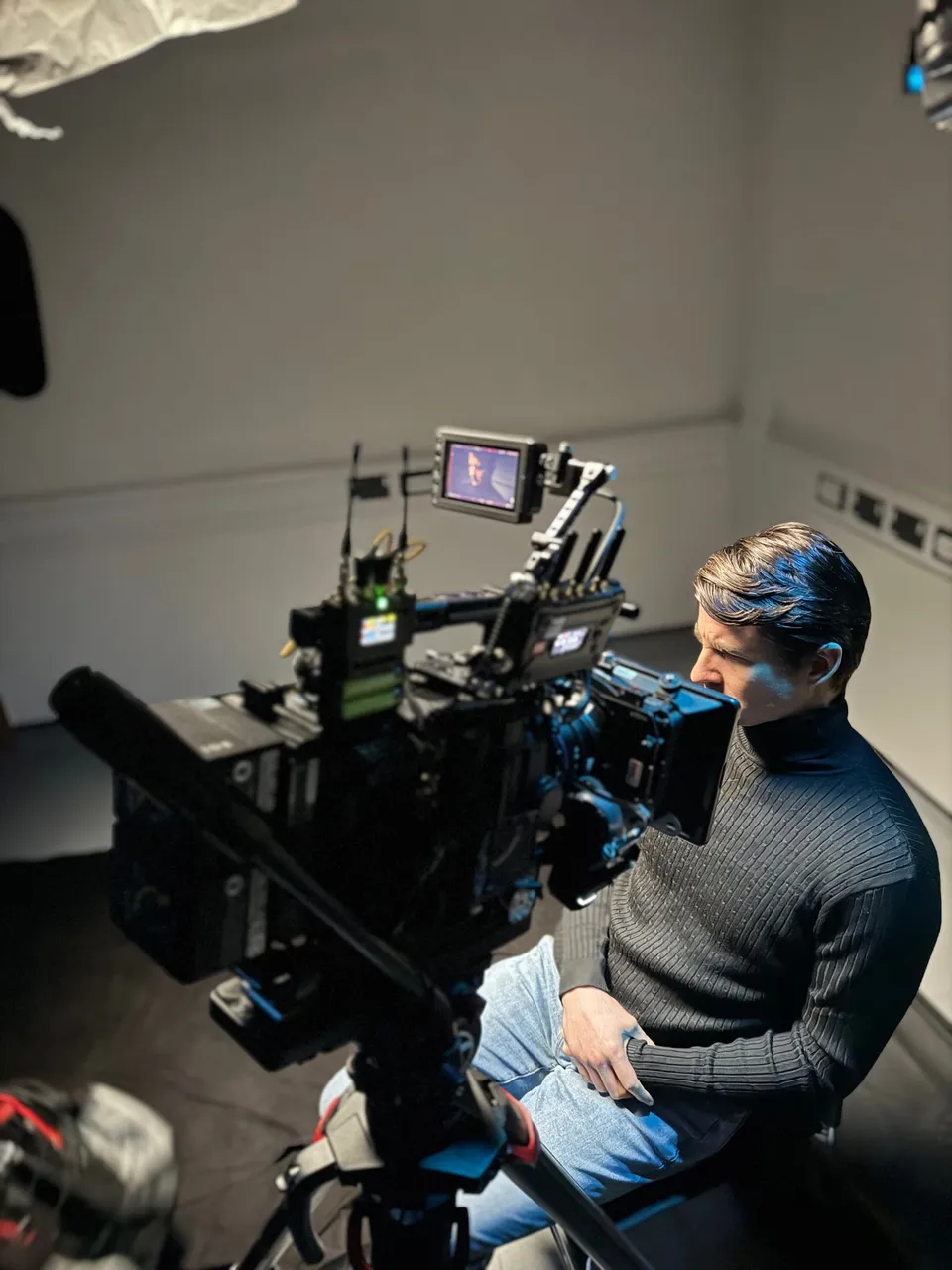 A man in a grey jumper sits beside a large cinema camera, looking towards its monitor, in a plain white room.