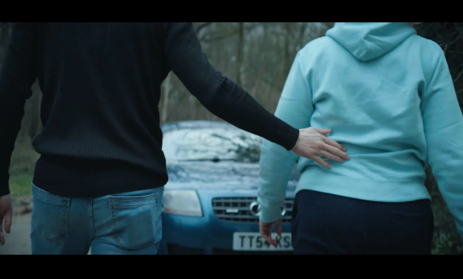 Close view from behind of two people walking towards a parked car, one resting a hand on the other's back, trees in the background.