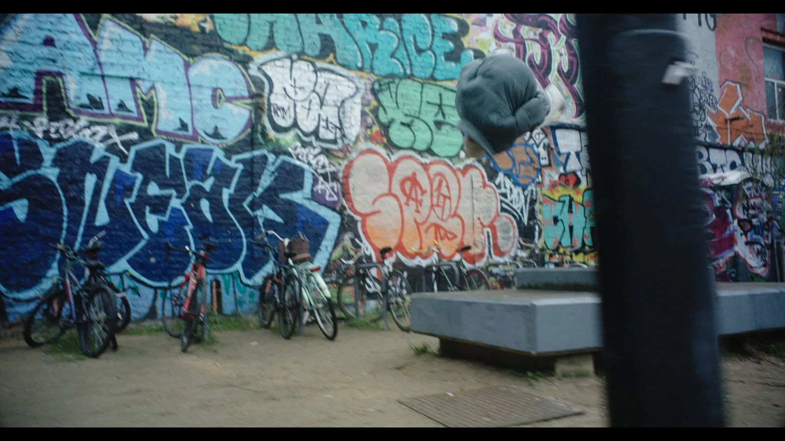 A blurred figure leaps in mid-air near a graffiti-covered wall, bicycles parked below and concrete blocks in the foreground.