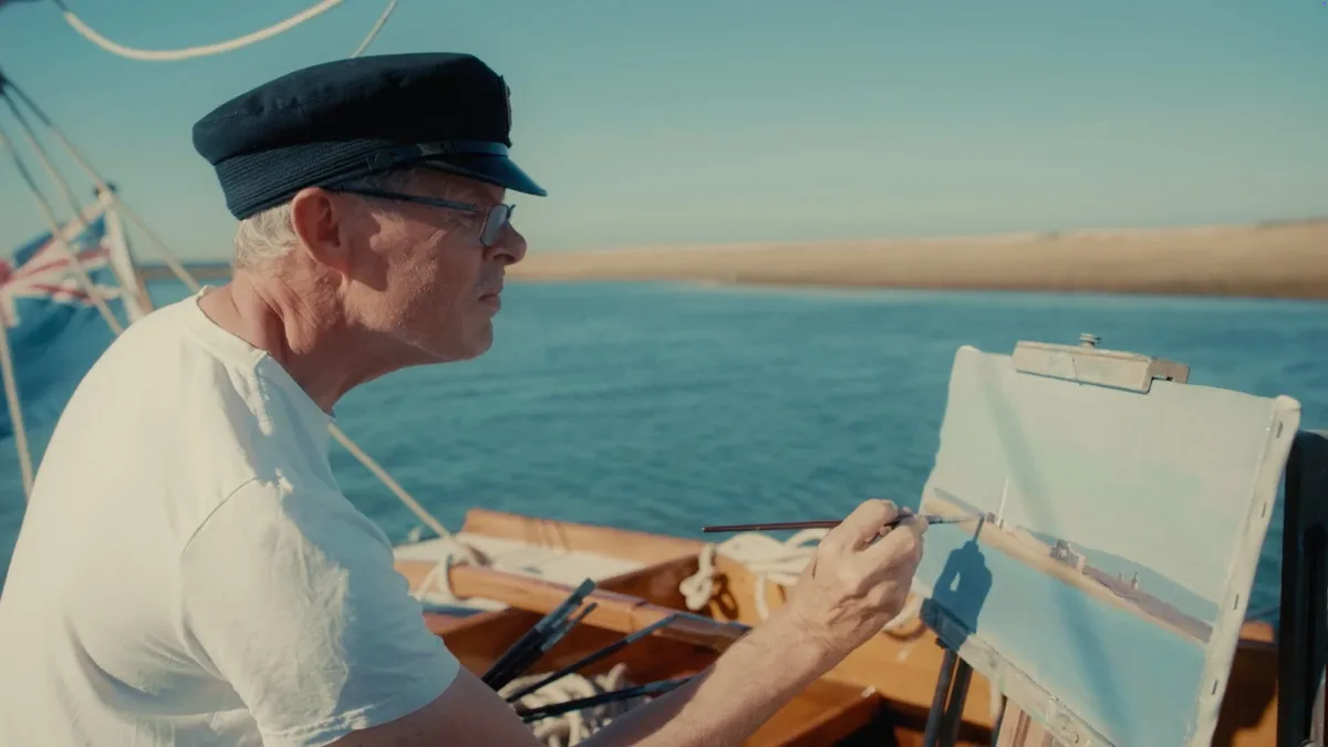 A grey-haired man in glasses and a dark peaked cap, in profile, paints at an easel aboard a boat, sandy shoreline and water behind him.