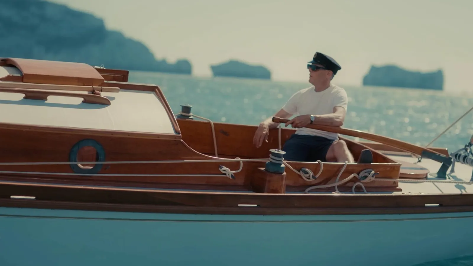 A man in a peaked cap and sunglasses, wearing a white T-shirt and dark shorts, steers a wooden motorboat, sea stacks visible in the distance.
