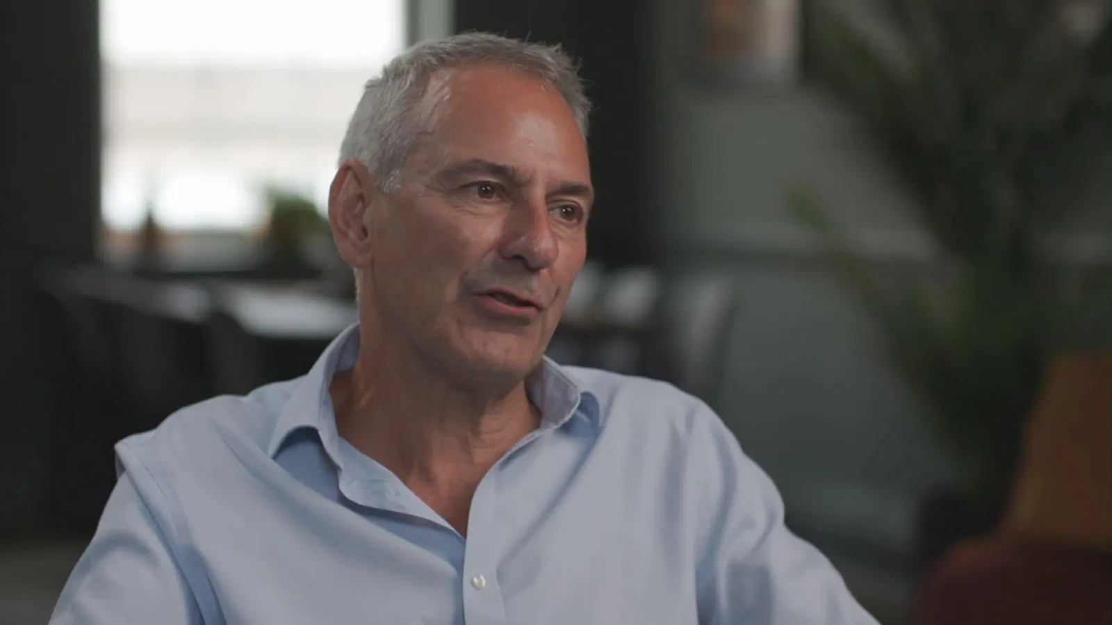 Close-up of a grey-haired man in a light blue shirt, speaking and looking off-camera, with a blurred plant and dark furniture behind him.
