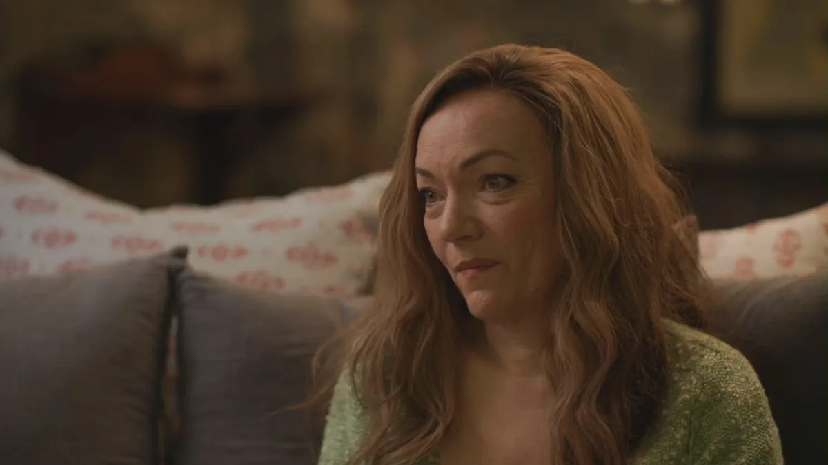 Close-up of a woman with long wavy reddish hair, in a green top, looking off-camera on a grey sofa with patterned cushions.