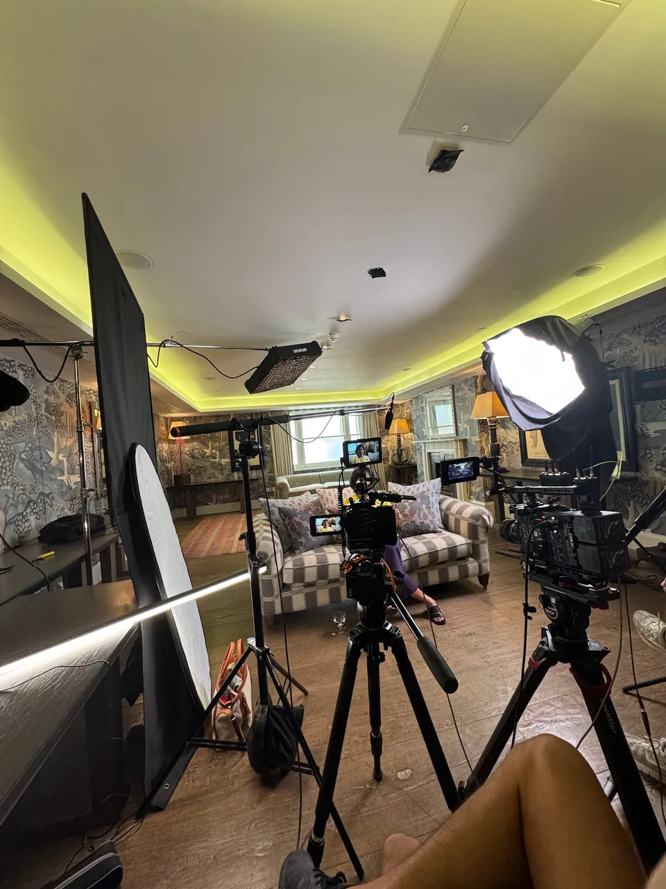 Camera crew filming a woman on a checked sofa in a patterned sitting room, with tripod-mounted cameras, monitors, softbox lights and a boom microphone in the foreground.