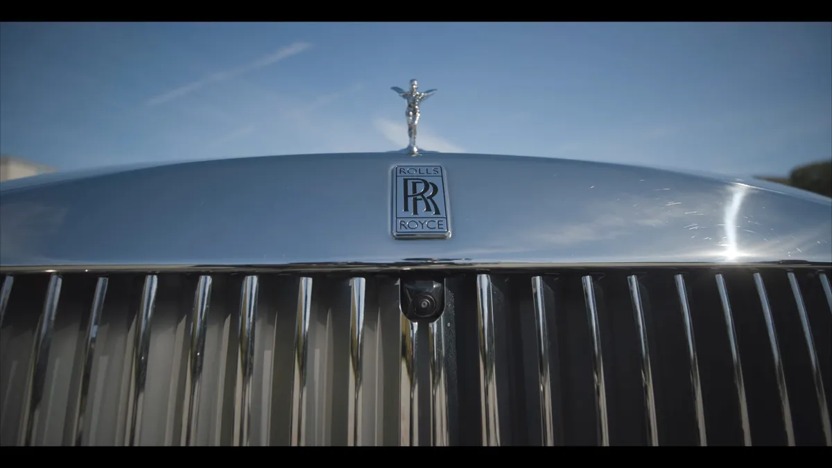 Close-up of a Rolls-Royce bonnet with the Spirit of Ecstasy mascot and badge above a chrome grille, set against a blue sky.