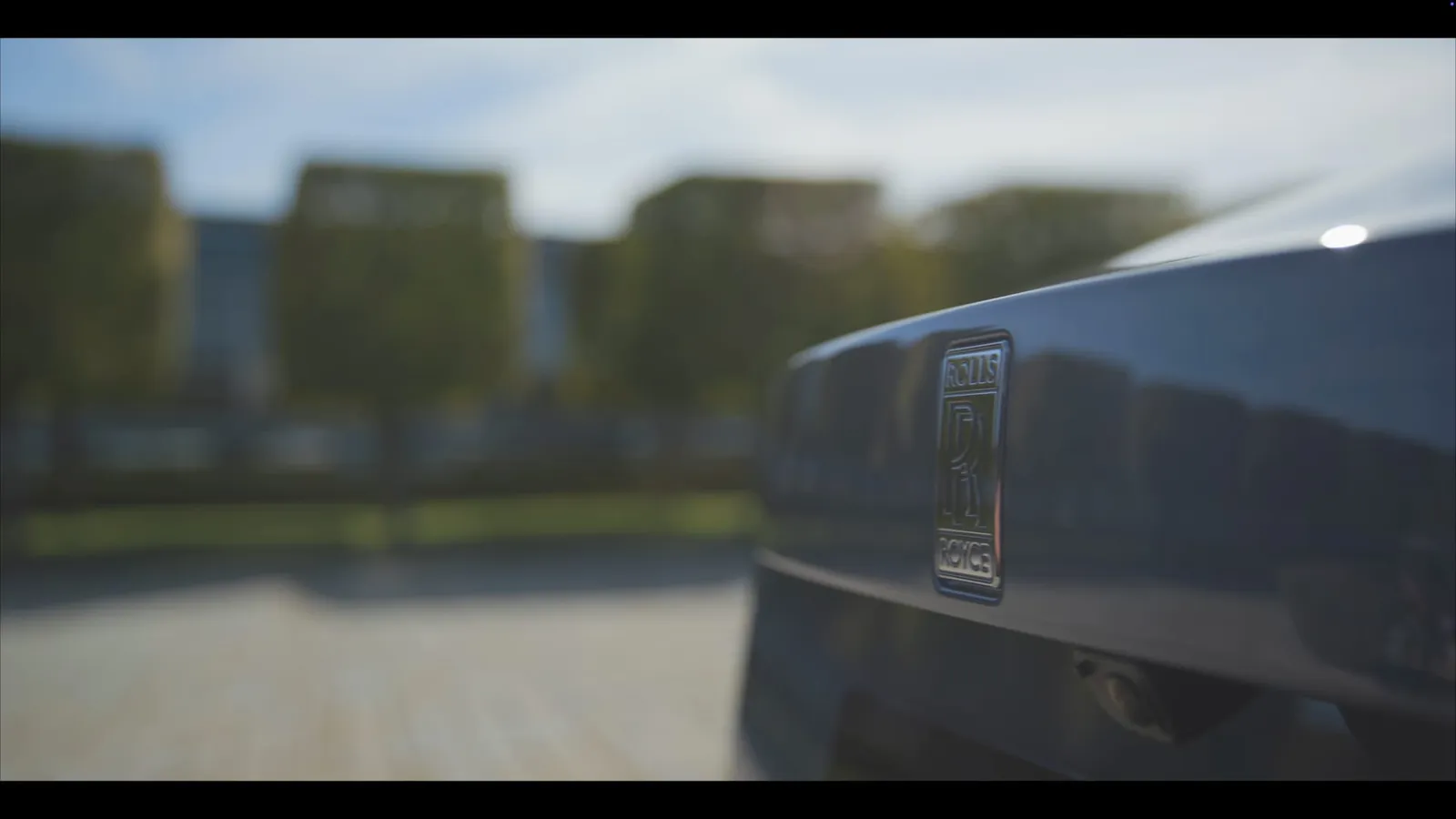 Close-up of a Rolls-Royce badge on a dark car's boot, with trees and buildings blurred in the background.