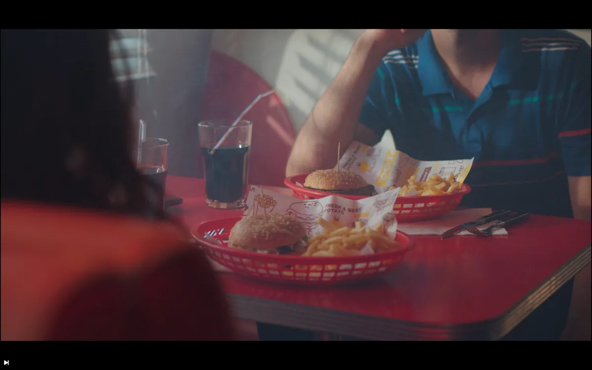 Two red plastic baskets of burgers and chips on a diner table, with soft drinks and a crew member seated beyond, out of focus in the foreground.