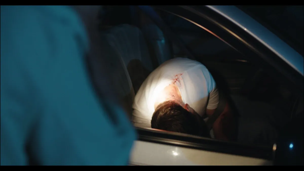 Torchlit close-up through a car window of a slumped figure in a bloodstained white t-shirt, a member's shoulder blurred in the foreground.