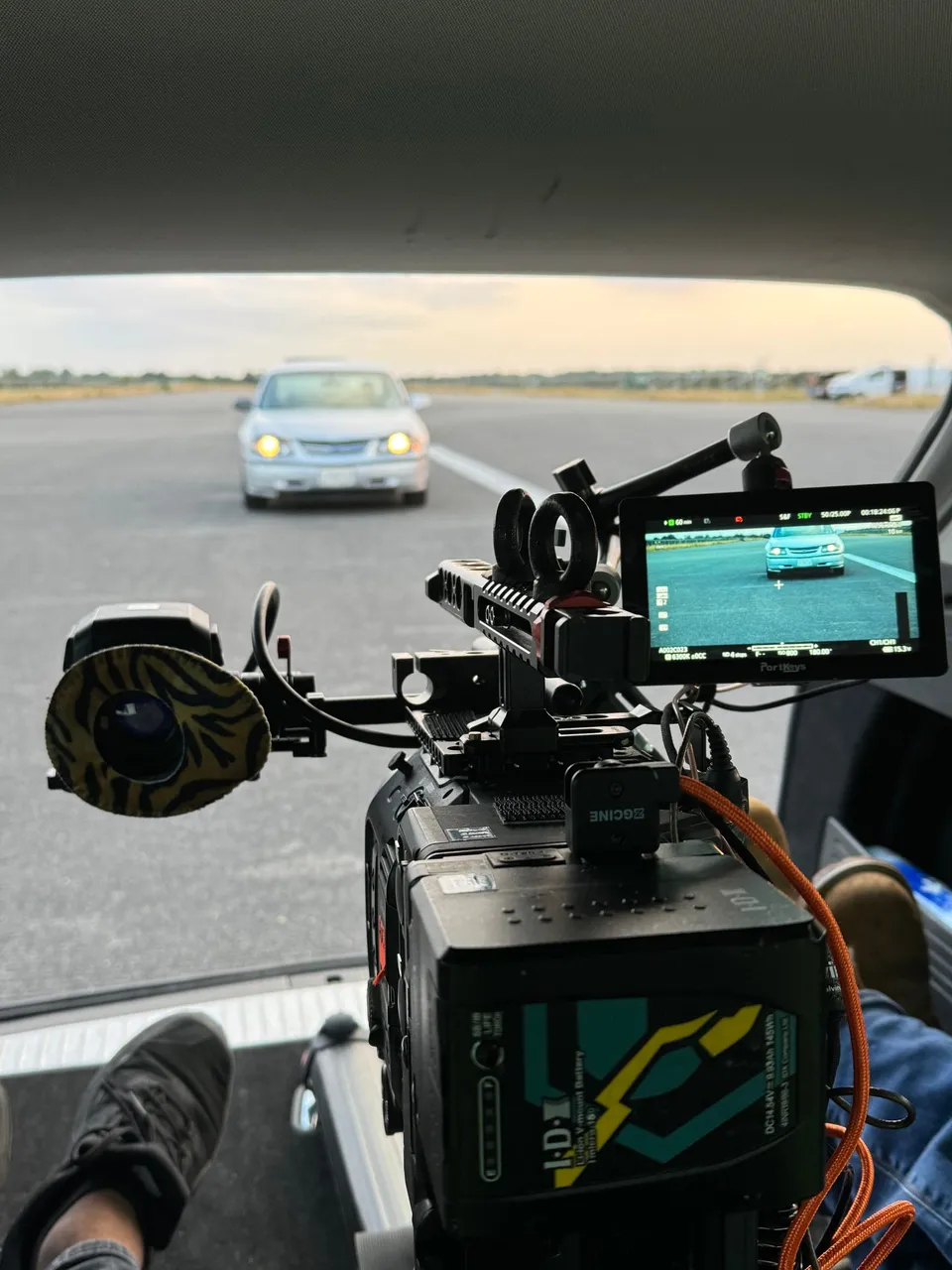 Camera rig with monitor mounted in a vehicle, filming a saloon car with headlights on approaching along an open road.